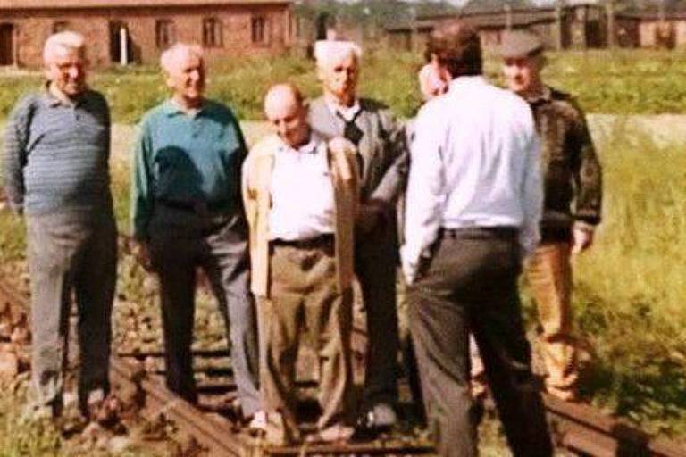 Um grupo de sete homens está posicionado sobre trilhos de trem. Eles estão vestidos de maneira casual， com algumas camisas de manga longa e suéteres. Ao fundo， há um edifício de tijolos e uma área verde. A expressão dos homens varia， alguns parecem pensativos. A cena sugere um momento de reflexão ou reunião.