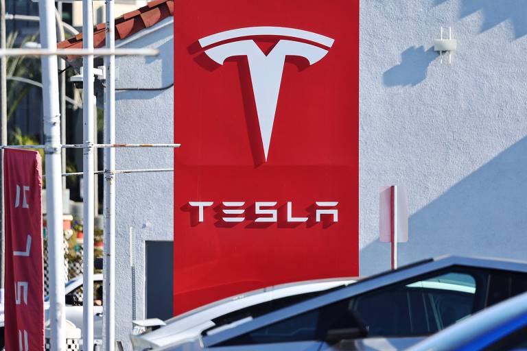 A imagem mostra um grande logotipo da Tesla em uma parede vermelha. O logotipo é um 039;T039; estilizado， e abaixo dele está escrito 039;TESLA039; em letras maiúsculas brancas. Ao fundo， é possível ver parte de um carro e outros elementos de um ambiente urbano.
