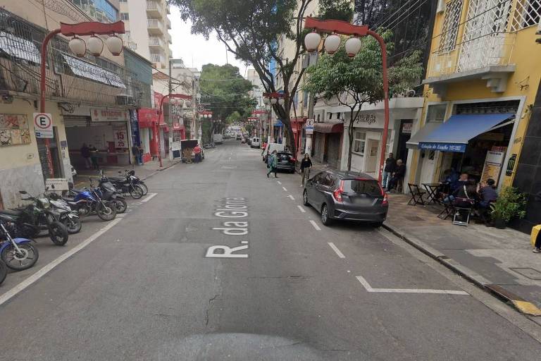 A imagem mostra a Rua da Glória em São Paulo， com uma via de mão dupla. À esquerda， há várias motos estacionadas e um comércio com letreiros visíveis. À direita， um carro está estacionado e algumas pessoas estão sentadas em mesas do lado de fora de um restaurante. A rua é arborizada， com árvores ao longo da calçada e postes de luz com lâmpadas. No fundo， é possível ver mais veículos e prédios