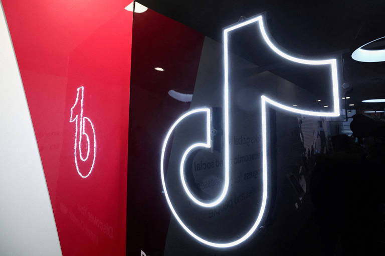 A imagem apresenta dois logos do TikTok. O logo maior， iluminado em branco， é o símbolo principal da plataforma， enquanto o logo menor， à esquerda， é uma versão simplificada em um fundo vermelho. O fundo da imagem é predominantemente preto.
