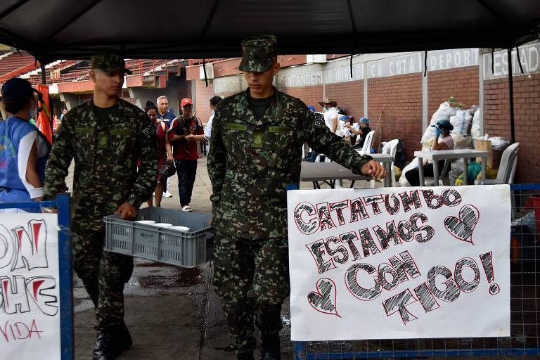 Dois soldados em uniforme militar， um segurando uma caixa e o outro segurando um cartaz que diz 039;CHETUMBO ESTAMOS CON TIGO!039;. Ao fundo， há pessoas e mesas com alimentos， sob uma estrutura coberta.