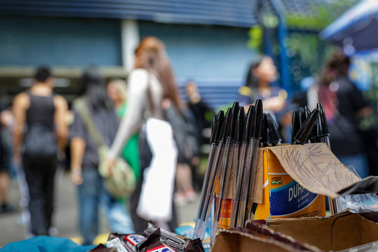 A imagem mostra uma banca de vendas com canetas em primeiro plano. As canetas estão organizadas em uma caixa de papelão. Ao fundo， há um grupo de pessoas em movimento， com algumas indistintas devido ao desfoque. O ambiente parece ser um espaço ao ar livre， possivelmente um mercado ou evento.