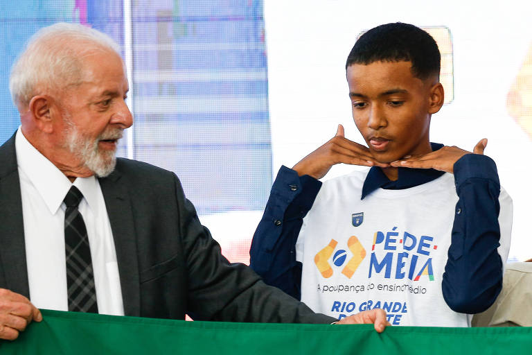 A imagem mostra um homem mais velho， com cabelo grisalho e barba， vestindo um terno escuro e gravata， ao lado de um jovem. O jovem está usando uma camiseta branca com o texto 039;Pé de Meia039; e faz uma pose com as mãos no rosto. Ambos estão em um evento， segurando uma faixa verde.
