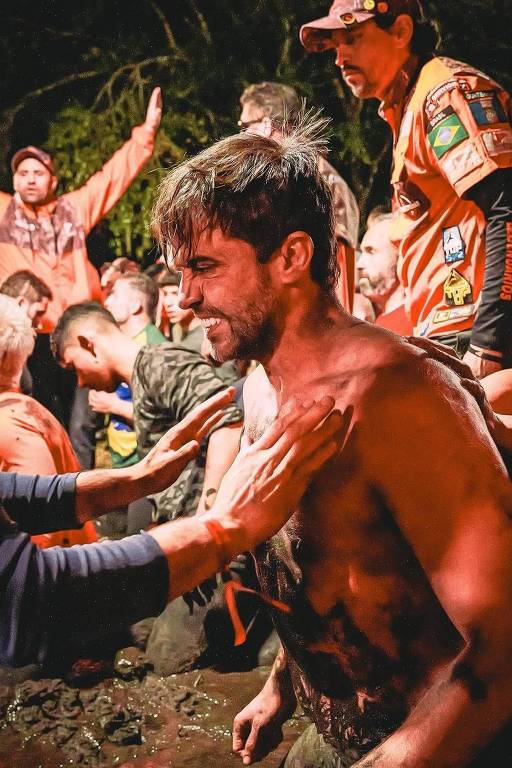 A imagem mostra um grupo de pessoas em um ambiente natural, possivelmente durante uma competição ou evento ao ar livre. Um homem sem camisa está em primeiro plano, com o corpo sujo de terra ou lama, enquanto outras pessoas ao fundo parecem estar animadas e participando do evento. Algumas pessoas estão vestindo camisetas de cores vibrantes, e há uma atmosfera de celebração e camaradagem.