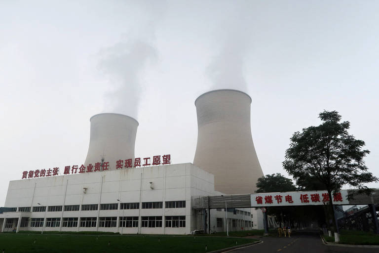 A imagem mostra uma usina nuclear com duas torres de resfriamento em destaque， emitindo vapor. Na parte inferior， há um edifício com uma fachada branca e uma inscrição em caracteres chineses. O céu está nublado， criando uma atmosfera de neblina.
