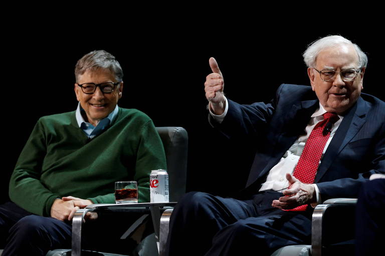 A imagem mostra Bill Gates e Warren Buffett sentados em cadeiras durante um evento. Gates está à esquerda， usando um suéter verde e óculos， com um copo e uma lata de refrigerante à sua frente. Buffett está à direita， vestindo um terno escuro e uma gravata vermelha， levantando o polegar em sinal de aprovação.
