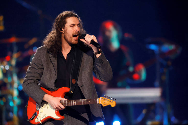 Um homem com cabelo longo e solto， vestido com um terno escuro， estaacute; cantando em um microfone enquanto toca uma guitarra eleacute;trica vermelha. Ao fundo， haacute; outros muacute;sicos e luzes coloridas iluminando o palco.