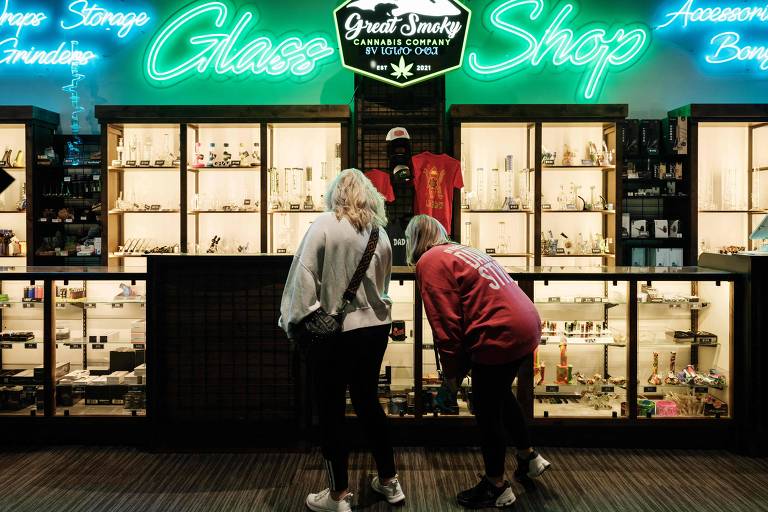 A imagem mostra duas pessoas em frente a uma vitrine de uma loja chamada 039;Great Smoky Glass Shop039;. A loja é iluminada com um letreiro neon verde que diz 039;Glass Shop039; e 039;Awesome Bongs039;. As prateleiras da vitrine exibem diversos produtos， incluindo bongs e outros acessórios relacionados ao uso de cannabis. As pessoas estão observando os itens expostos.
