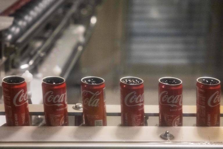 A imagem mostra uma linha de produção com várias latas de refrigerante da marca Coca-Cola. As latas estão dispostas em uma superfície metálica， alinhadas uma ao lado da outra， com a parte superior visível. O fundo é desfocado， sugerindo um ambiente industrial.