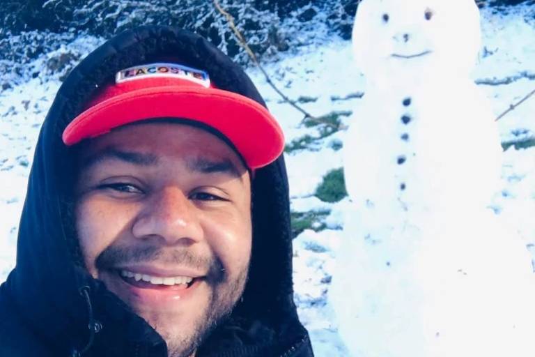 Um homem sorridente tira uma selfie em um ambiente coberto de neve. Ele está usando um casaco preto e um boné vermelho com a marca Lacoste. Ao fundo, há um boneco de neve com um sorriso, feito de neve, com botões pretos e um pouco de grama no topo.