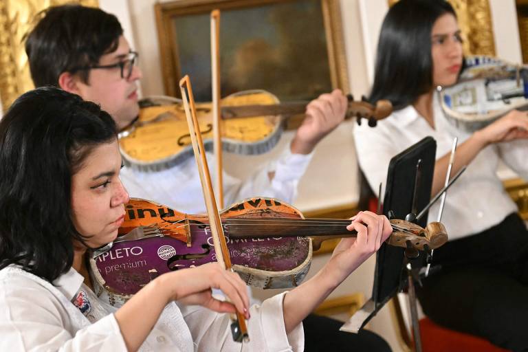 A imagem mostra três músicos tocando violinos em um ambiente elegante. A primeira musicista, à esquerda, tem cabelo preto e usa uma camisa branca, segurando um violino decorado com rótulos coloridos. O músico ao centro, com óculos, também toca um violino semelhante. À direita, uma musicista com cabelo liso e escuro toca seu violino, que também apresenta uma decoração peculiar. O fundo é composto por uma moldura dourada e uma pintura.