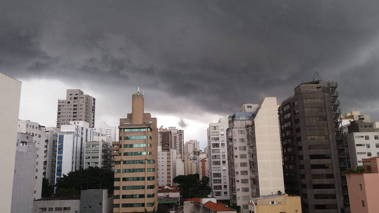 A imagem mostra uma vista urbana com prédios altos e um céu escuro e nublado. As nuvens são densas e ameaçadoras， sugerindo a possibilidade de chuva. A cidade é composta por uma variedade de edifícios， incluindo alguns de vidro e outros de concreto， com áreas verdes visíveis entre os prédios.