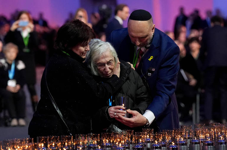 A imagem mostra três pessoas em uma cerimônia. Duas mulheres estão abraçando uma mulher idosa， que parece emocionada. Um homem， vestido com um terno escuro e um kippah， está ajudando a idosa com uma vela. Há várias velas acesas e uma multidão de pessoas assistindo ao evento.