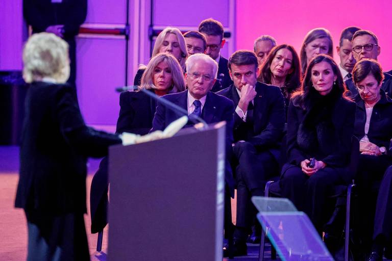 A imagem mostra um evento onde uma mulher está falando em um púlpito. No fundo， há um grupo de pessoas sentadas， incluindo um homem com cabelo grisalho e outros indivíduos em trajes escuros. A iluminação do ambiente é em tons de roxo e azul， criando uma atmosfera formal.