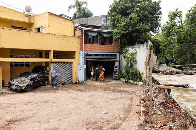 A imagem mostra uma área externa de uma casa com uma garagem. À esquerda， há um carro danificado， aparentemente abandonado， com a parte frontal amassada. A garagem está fechada com uma porta de metal. Algumas pessoas estão em pé na entrada da garagem， enquanto outras estão próximas a um espaço vazio ao fundo. O ambiente é cercado por vegetação e há uma construção em ruínas à direita， com terra exposta e detritos visíveis. O céu está parcialmente nublado.