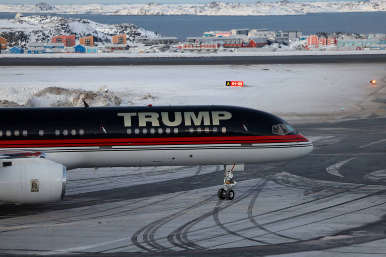 Um avião comercial com a inscrição 039;TRUMP039; na fuselagem está estacionado em uma pista coberta de gelo. O avião possui uma pintura preta com detalhes em vermelho e uma bandeira dos Estados Unidos na cauda. Ao fundo， há uma paisagem de cidade com edifícios e uma área de água， sob um céu em tons de rosa e azul.