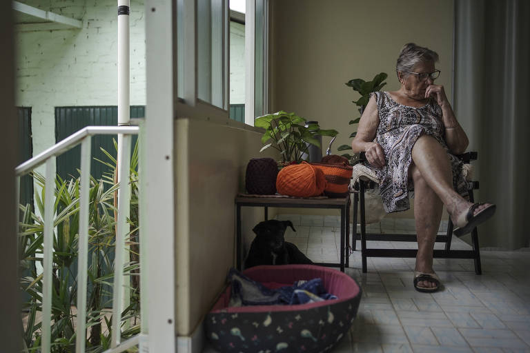 Uma mulher idosa está sentada em uma cadeira em uma varanda. Ela usa um vestido estampado e está com uma expressão pensativa. Ao seu lado， há uma mesa com plantas e objetos pessoais， além de um cesto para animais de estimação no chão. A varanda é iluminada e tem uma vista para um espaço verde.