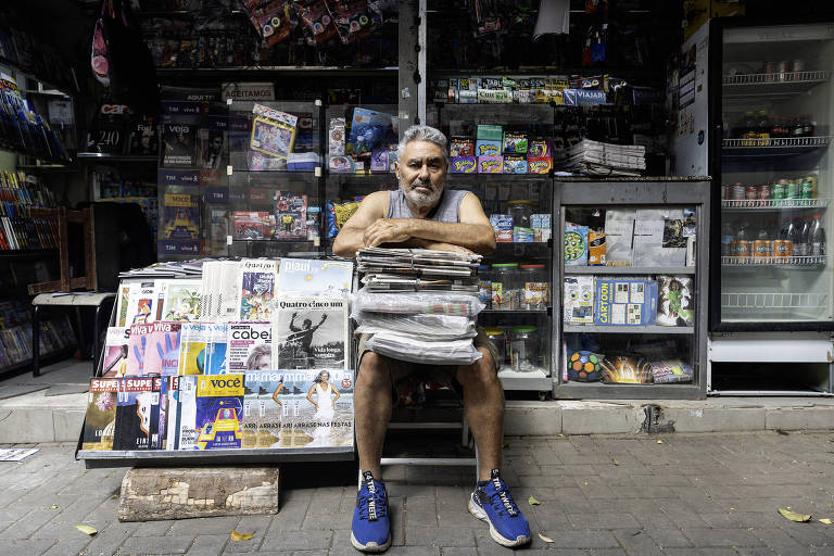 Um homem sentado em uma banca de jornal, com uma pilha de jornais e revistas à sua frente. Ele está usando uma camiseta e shorts, com tênis azuis. Ao fundo, a banca está cheia de produtos, incluindo bebidas e outros itens à venda.