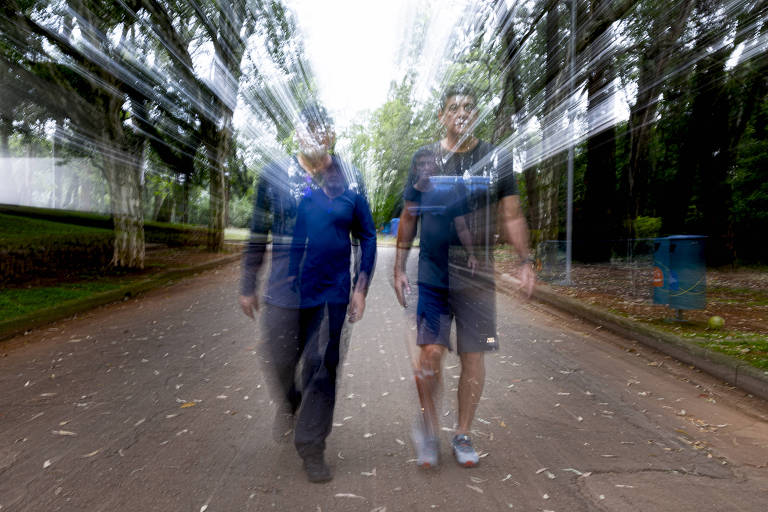 A imagem mostra duas pessoas caminhando em um caminho de parque. A foto está desfocada， criando um efeito de movimento. Ao fundo， há árvores e um espaço verde， com um banco visível à esquerda e um recipiente azul à direita.