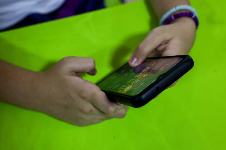 A imagem mostra as mãos de uma pessoa segurando um smartphone. O dispositivo está virado para baixo， exibindo uma tela com um jogo ou aplicativo. A superfície em que as mãos estão apoiadas é de cor verde brilhante.