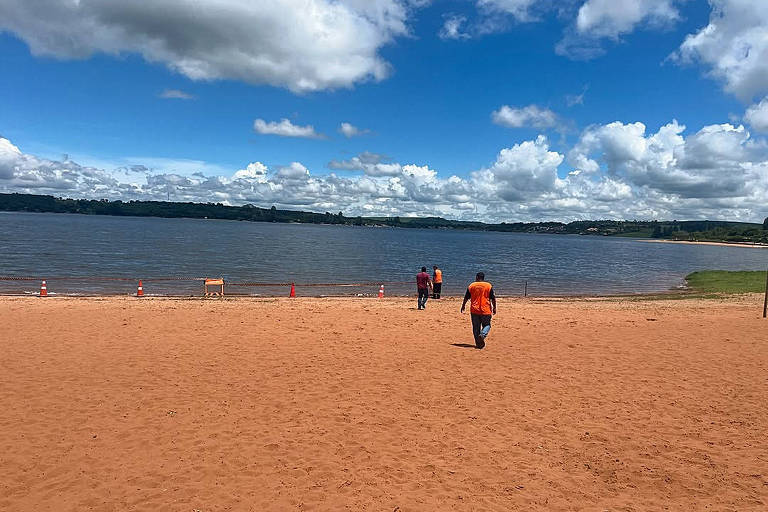 A imagem mostra uma praia de areia clara com algumas pessoas caminhando em direção à água. O céu está azul com nuvens brancas e volumosas. Ao fundo， é possível ver um lago e vegetação nas margens.