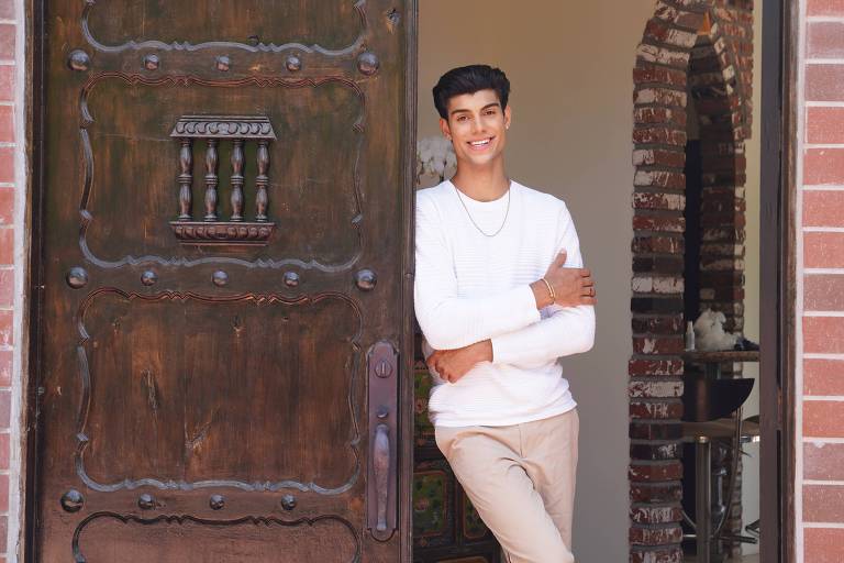 Um jovem está posando na entrada de uma casa， apoiado na porta de madeira. Ele usa uma camiseta branca de manga longa e calças claras. O fundo mostra um interior com paredes de tijolos e uma decoração visível. A porta tem detalhes ornamentais e uma janela de vidro na parte superior.