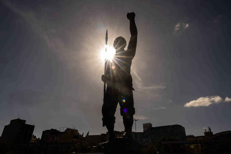 A imagem mostra uma estátua de um homem em posição de luta， levantando um braço e segurando uma lança. O sol brilha atrás da estátua， criando um efeito de silhueta. O céu está parcialmente nublado， com algumas nuvens visíveis.
