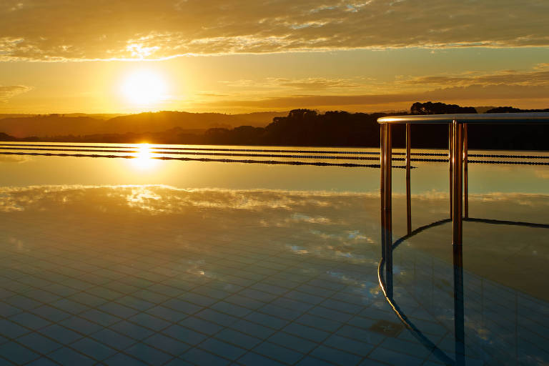 A imagem mostra uma piscina com borda infinita refletindo o céu ao entardecer. O sol está se pondo no horizonte， criando um efeito dourado na água. Nuvens estão dispersas no céu， e há uma escada de metal na borda da piscina.