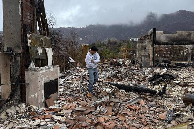 A imagem mostra um jovem em meio a escombros de uma construção destruída por um incêndio. O chão está coberto de destroços， incluindo pedaços de telhas e materiais de construção. Ao fundo， há uma estrutura parcialmente queimada e uma paisagem montanhosa com nuvens escuras. O céu está nublado， indicando um clima sombrio.