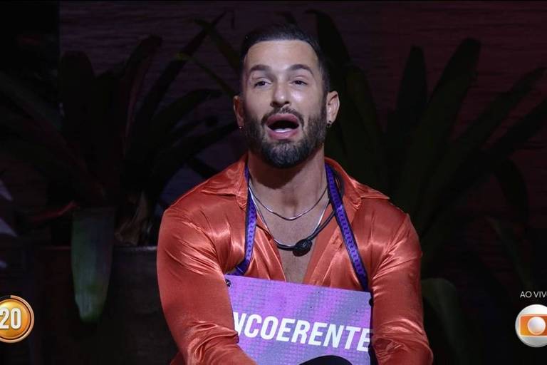 Um homem com cabelo curto e barba está sentado em um ambiente iluminado, usando uma camisa laranja brilhante. Ele segura uma placa com a palavra 'INCOERENTE' escrita em letras grandes e roxas. Ao fundo, há plantas e uma tela que indica que é uma transmissão ao vivo.