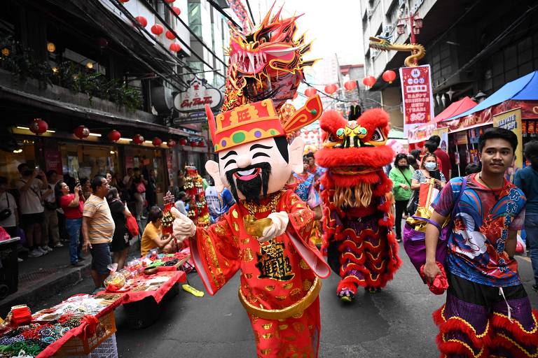 A imagem mostra um desfile de festival em uma rua movimentada, com pessoas vestindo trajes coloridos. Um personagem grande e sorridente, vestido de vermelho e com um chapéu decorado, lidera o grupo. Atrás dele, há um dragão dançante e outras pessoas em trajes festivos. O ambiente é decorado com lanternas vermelhas e há barracas de comida e produtos ao longo da rua.