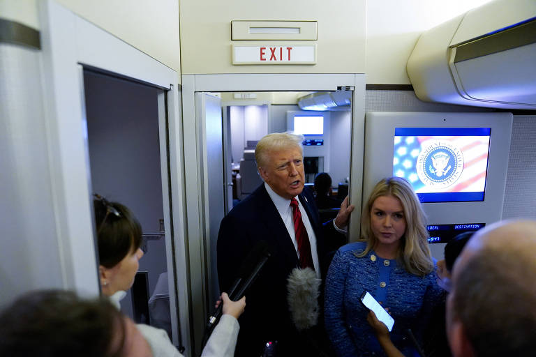 A imagem mostra um grupo de pessoas em um espaço confinado， possivelmente dentro de um avião. Um homem de cabelo loiro e terno escuro está falando， enquanto uma mulher ao seu lado segura um celular. Outras pessoas estão com microfones e gravadores， fazendo perguntas. Ao fundo， há uma tela com o emblema presidencial e uma bandeira dos Estados Unidos. Uma placa acima da porta indica a saída.