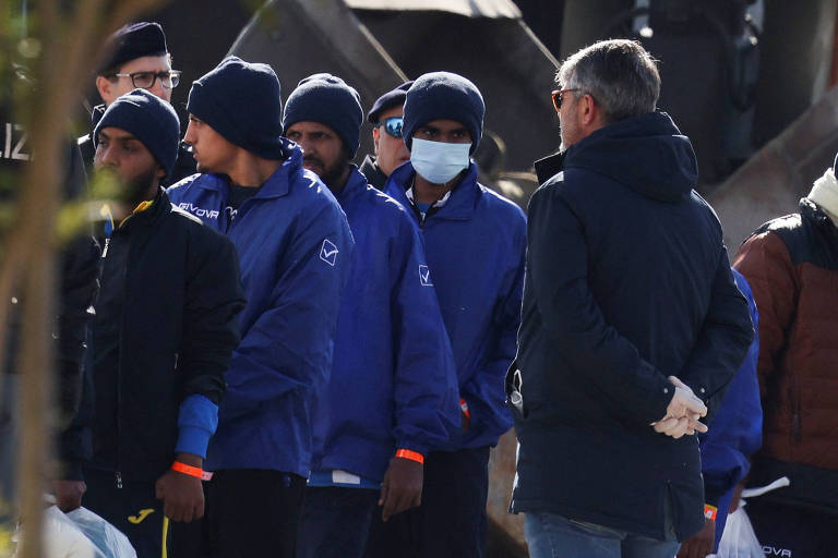 Um grupo de homens está em pé em um ambiente urbano. Eles estão vestidos com jaquetas azuis e gorros， alguns usando máscaras faciais. Um homem de costas， vestido com um casaco escuro， está conversando com eles. O cenário parece ser ao ar livre， com uma estrutura metálica ao fundo.