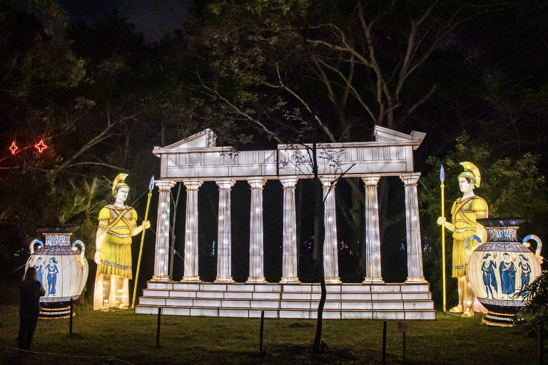 A imagem mostra uma instalação de luzes noturna representando uma estrutura arquitetônica com colunas, semelhante a um templo grego. À esquerda, há uma figura iluminada de um guerreiro com armadura e lança, e à direita, uma figura semelhante segurando um escudo. Também há um grande vaso decorado com figuras humanas. O fundo é escuro, com árvores e algumas luzes ao longe.