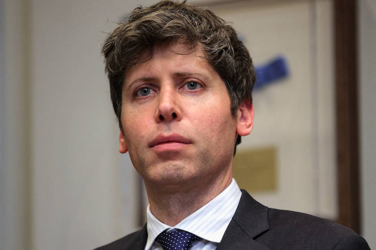 Imagem mostra Sam Altman， um homem com cabelo castanho claro e liso， usando um terno escuro e uma gravata azul. Ele está em pé， com uma expressão séria， em um ambiente interno que parece ser um escritório. Ao fundo， há uma parede com uma obra de arte e um objeto azul visível.