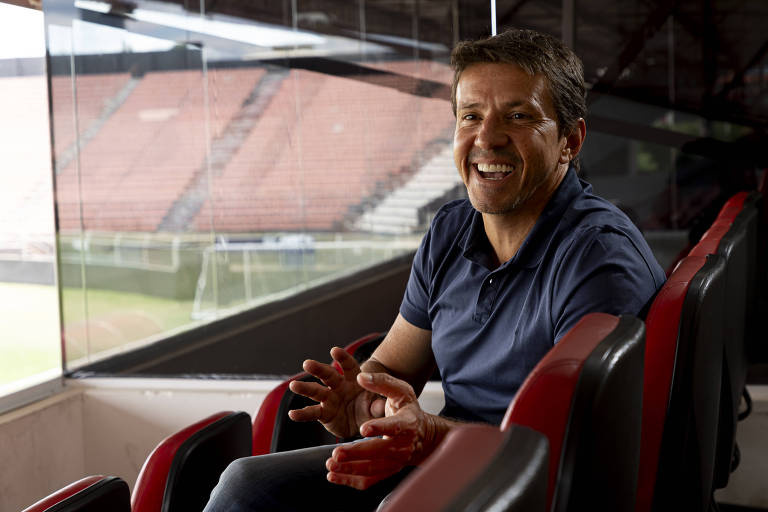 Um homem sorridente está sentado em uma cadeira de estádio， com as mãos gesticulando. Ele usa uma camisa polo azul e está em um ambiente que parece ser uma cabine de imprensa ou um espaço reservado em um estádio de futebol. Ao fundo， é possível ver as arquibancadas vazias e o campo de futebol.
