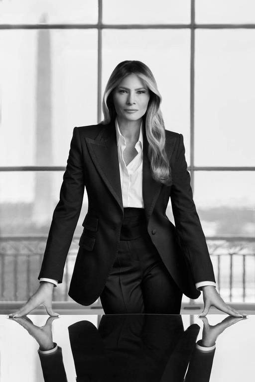 A imagem mostra uma mulher em um traje formal preto， com uma camisa branca por baixo. Ela está em pé， apoiando as mãos sobre uma mesa de vidro， com uma expressão séria. Ao fundo， há grandes janelas que permitem a entrada de luz， e é possível ver um monumento ao longe， sugerindo que a cena se passa em um ambiente urbano. A imagem é em preto e branco， destacando os contrastes.