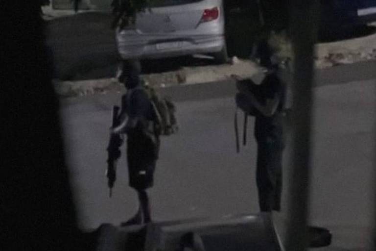 A imagem mostra duas pessoas caminhando na calçada de uma rua. Ao fundo， há árvores e um veículo estacionado. A cena é observada através de uma abertura， possivelmente uma janela， que cria um efeito de sombra ao redor.