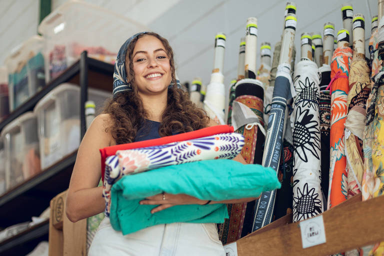 Uma mulher sorridente está em uma loja de tecidos， segurando três pilhas de tecidos coloridos. Ao fundo， há prateleiras com rolos de tecidos variados e caixas organizadas. A mulher usa uma blusa sem mangas e calças claras.