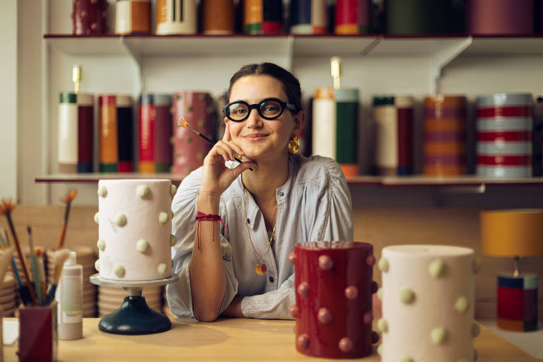 Uma mulher com óculos está sentada em uma mesa， sorrindo e apoiando o queixo na mão. Ao fundo， há várias latas coloridas dispostas em prateleiras. Na mesa， há dois recipientes decorativos， um branco com bolinhas e outro vermelho. O ambiente é bem iluminado e transmite uma sensação criativa.