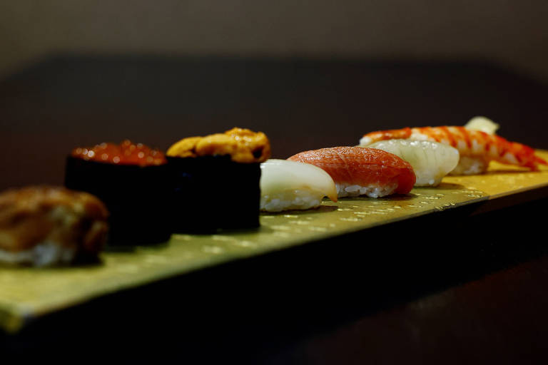 A imagem mostra um prato de sushi disposto em uma tábua. Há diferentes tipos de sushi， incluindo nigiri com peixe， sushi enrolado com alga e recheio， e camarão. O fundo é escuro， destacando os alimentos.