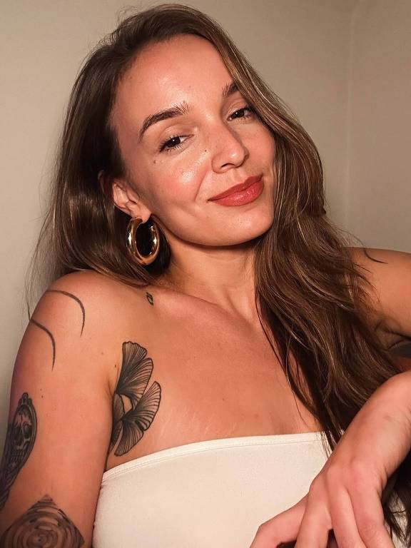 A imagem mostra uma mulher com cabelo longo e ondulado, usando um top branco. Ela está sorrindo levemente e olhando para a câmera. A mulher tem brincos grandes e dourados, e apresenta tatuagens visíveis em seu braço esquerdo, incluindo uma borboleta e um desenho em forma de olho. O fundo é de cor clara, e a iluminação é suave.