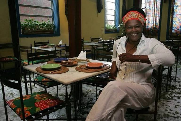 Uma pessoa sorridente sentada à mesa de um restaurante. Ela usa uma blusa branca e um lenço colorido na cabeça. Na mesa， há pratos coloridos e talheres. O ambiente é acolhedor， com mesas e cadeiras de metal， e janelas que permitem a entrada de luz natural.