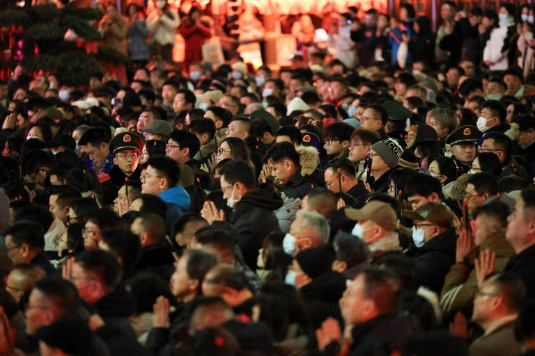 A imagem mostra uma grande multidão reunida em um evento noturno. As pessoas estão usando máscaras e algumas estão com as mãos unidas em um gesto de oração ou respeito. O ambiente é iluminado por luzes quentes， criando uma atmosfera de celebração ou reflexão. A multidão é densa， com rostos visíveis em diferentes ângulos.