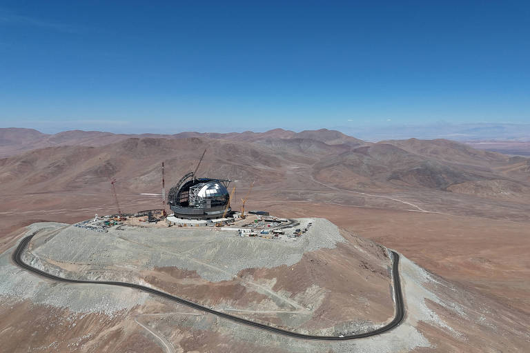 A imagem mostra um observatório astronômico localizado em uma área montanhosa e árida. O observatório possui uma estrutura grande e arredondada， com uma cúpula， e está situado no topo de uma elevação. Ao redor， há um terreno desértico com montanhas ao fundo e um céu azul claro.