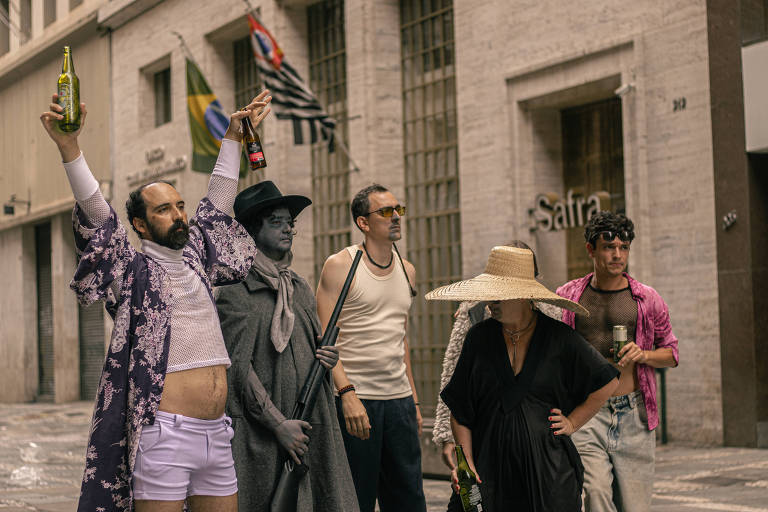 Um grupo de cinco pessoas se destaca em uma rua, cada uma vestindo roupas excêntricas. Um homem à esquerda levanta duas garrafas, usando uma camisa de manga longa com estampa floral e shorts brancos. Ao seu lado, um homem pintado de cinza, vestido com um manto e chapéu, segura um objeto que parece ser uma arma. Um terceiro homem, com óculos escuros e camiseta sem mangas, observa. Uma mulher usa um vestido preto e um chapéu de palha grande, enquanto um homem à direita, com cabelo encaracolado, segura uma lata de bebida e veste uma camisa rosa transparente. Ao fundo, bandeiras do Brasil e de São Paulo são visíveis.