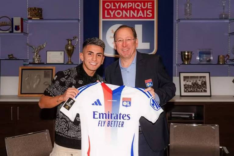 Na imagem， um jovem jogador e um homem mais velho estão sorrindo enquanto seguram uma camisa do Olympique Lyonnais. O jogador está à esquerda， vestindo uma camisa preta， e o homem à direita está usando um blazer azul. Ao fundo， há prêmios e troféus expostos em prateleiras， além de um logotipo do clube na parede.