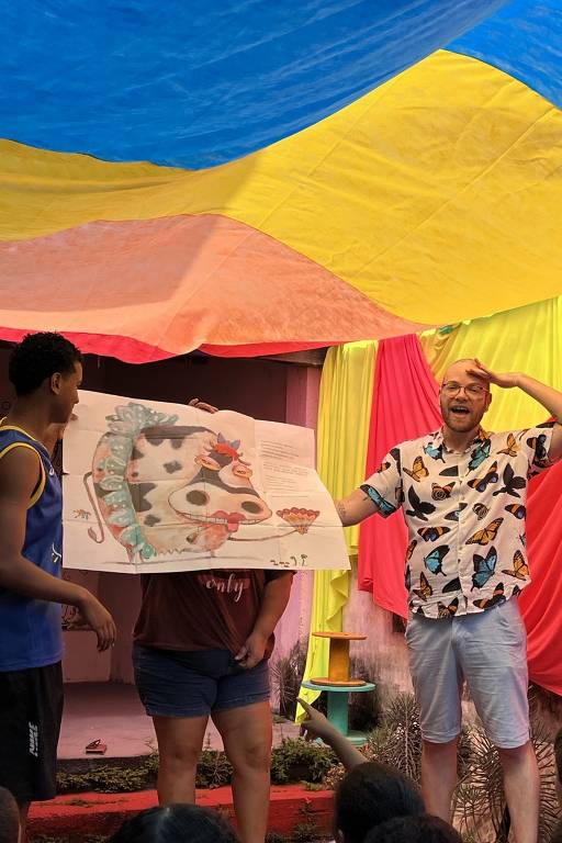 A imagem mostra um grupo de três pessoas em uma apresentação. Um homem de camisa colorida com estampas de borboletas está à direita, sorrindo e gesticulando. No centro, uma mulher segura um livro ilustrado com uma vaca e outros elementos coloridos, enquanto um jovem à esquerda observa. O fundo é decorado com tecidos coloridos, criando um ambiente festivo. Algumas crianças podem ser vistas na parte inferior da imagem, assistindo à apresentação.