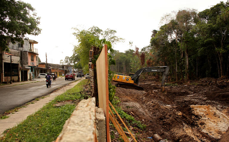 A imagem mostra um canteiro de obras em uma área urbana. À esquerda， há uma rua com uma motocicleta e um carro passando. À direita， o terreno está desmatelado e há uma máquina de construção， possivelmente uma escavadeira， trabalhando. Ao fundo， vê-se uma construção e árvores ao redor.