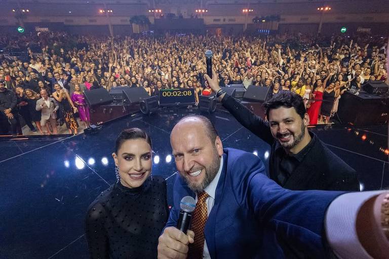 A imagem mostra um grupo de três pessoas tirando uma selfie em um evento ao vivo. No fundo, há uma grande multidão de pessoas assistindo ao evento. As duas pessoas à esquerda e à direita do homem no centro estão sorrindo. O homem no centro segura um microfone e está vestido com um terno azul e uma gravata laranja. O ambiente parece ser um palco iluminado.